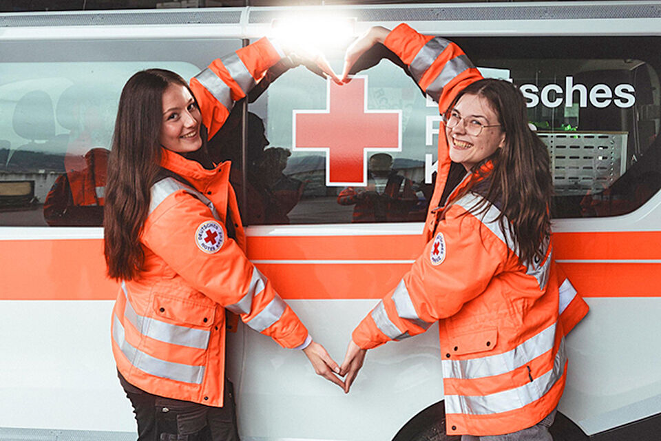 zwei DRklerinnen zeigen Herz vor einem Rettungswagen